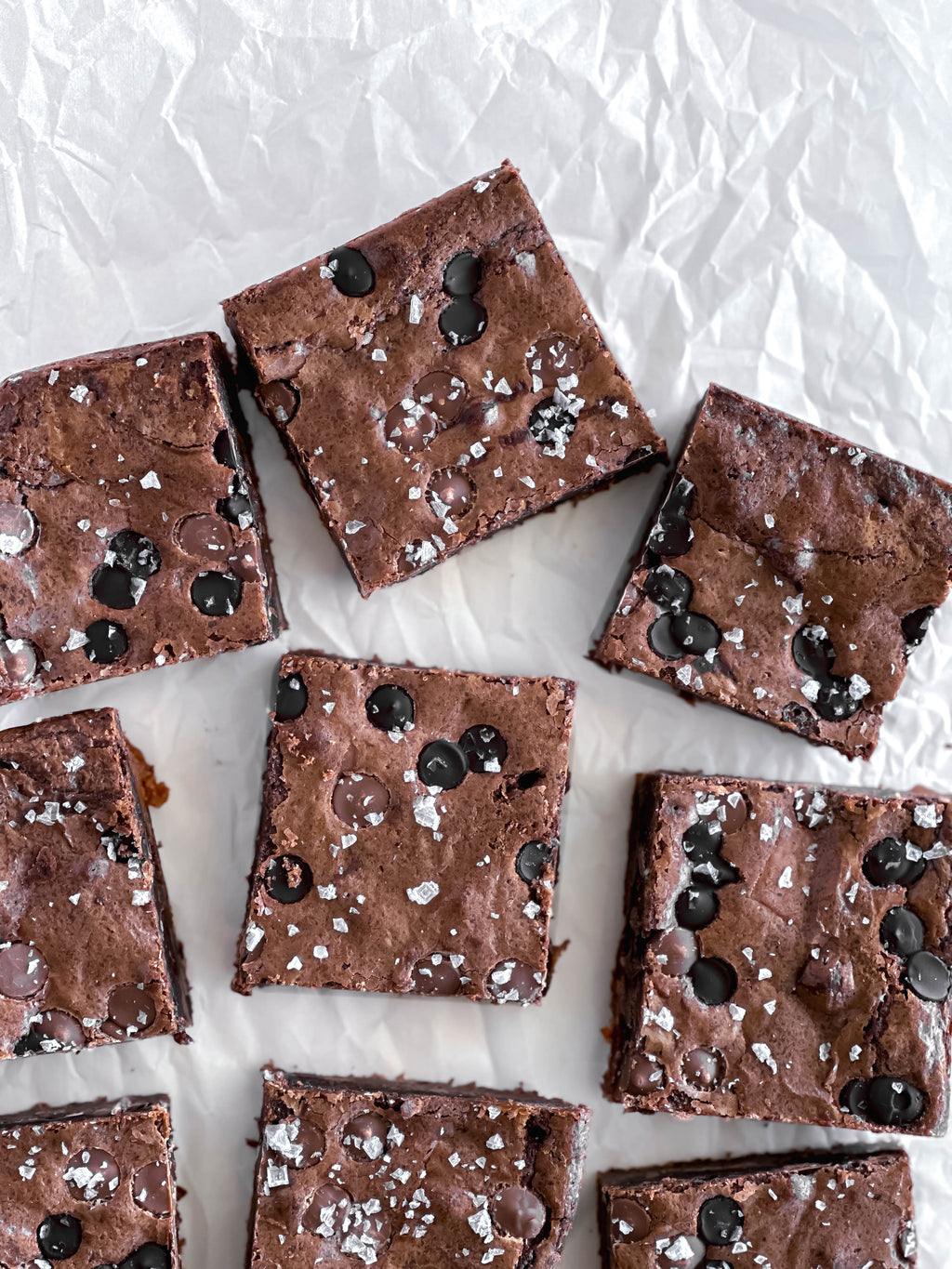 Chocolate brownies with chocolate chips and flaky sea salt on a white surface.