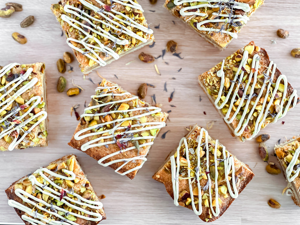Honey pistachio dessert bars with white chocolate drizzle and pistachios on a wooden surface.