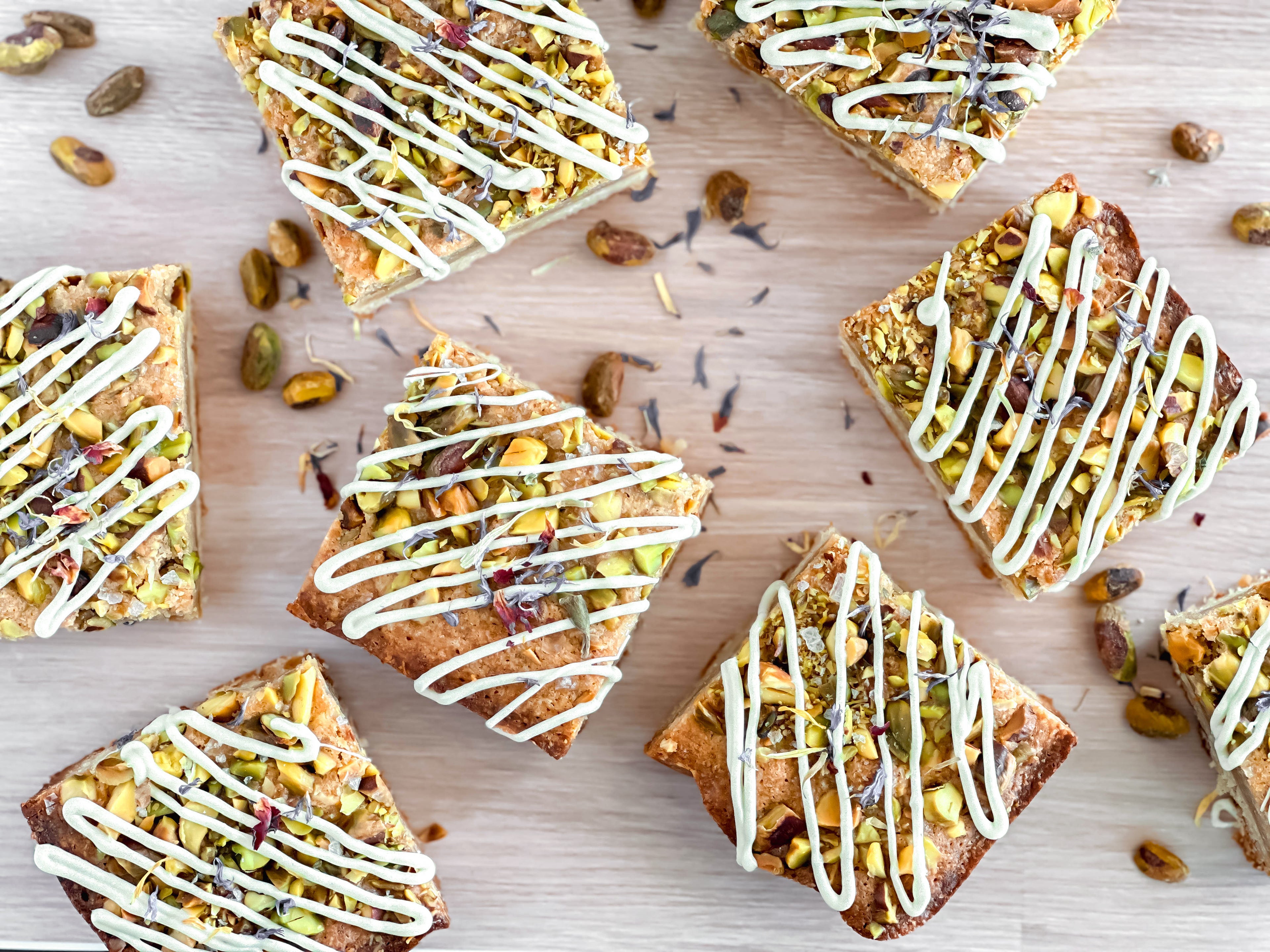 Honey pistachio dessert bars with white chocolate drizzle and pistachios on a wooden surface.