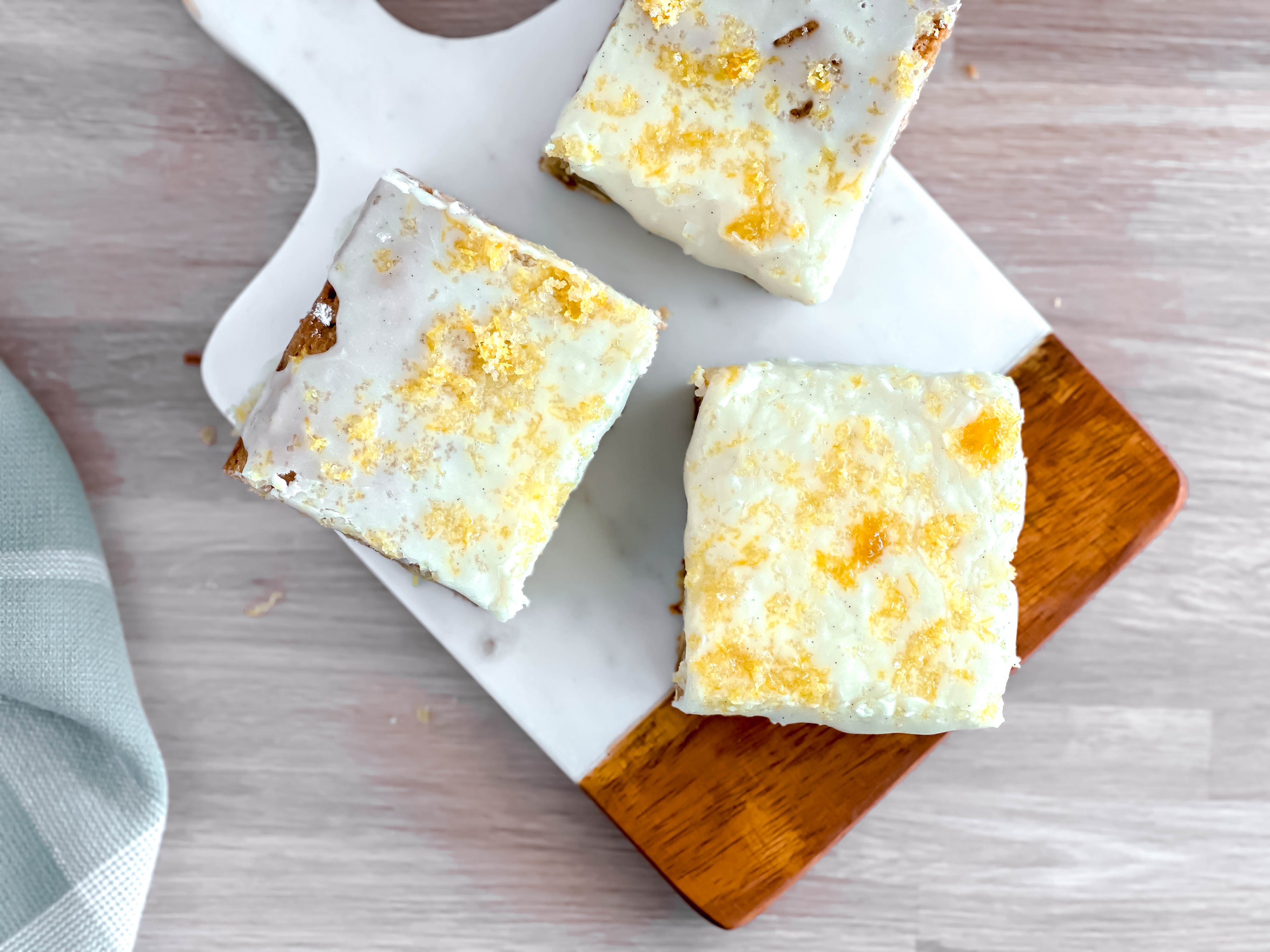 Three lemon blondies on a wooden and marble board, topped with sugar glaze, sprinkled with sugar and lemon zest.