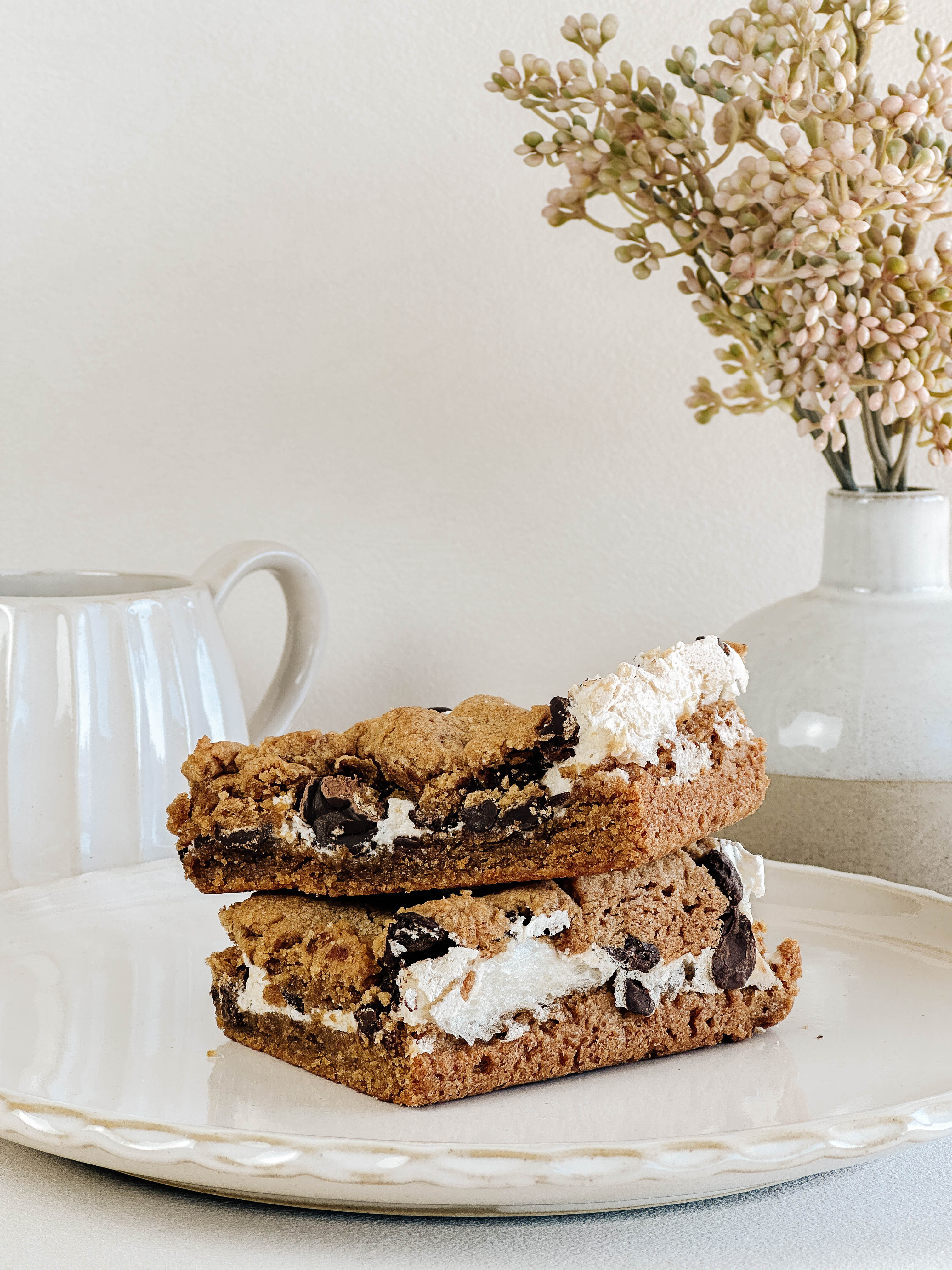 Two peanut butter s'mores bars stacked on a plate with a vase of flowers in the background.
